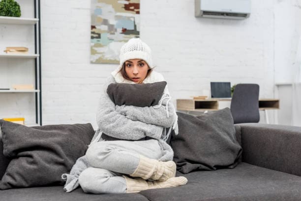 A woman holding a couch cushion inside her house while wearing warm clothes sitting on the couch.