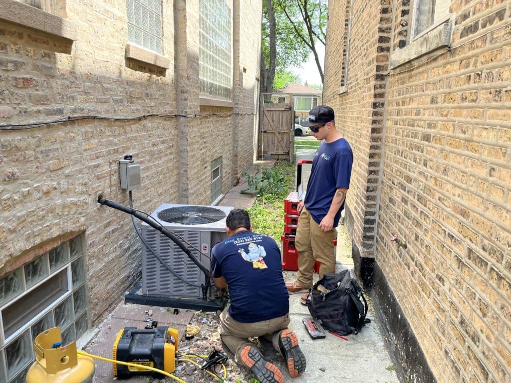 Two technicians working on an AC condenser unit in a gangway.